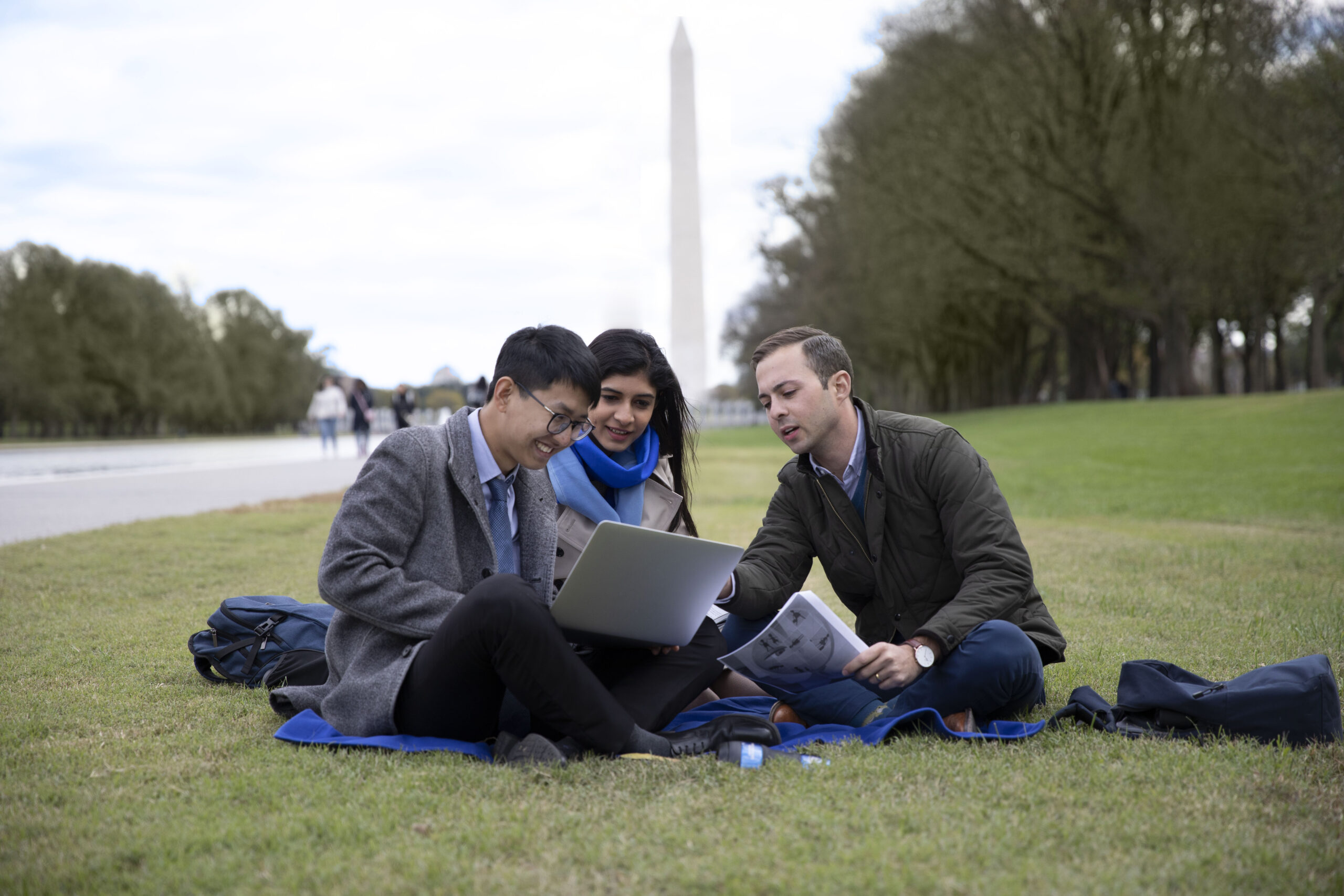 Three students look at a laptop while sitting in the grass; the Washington Monument is visible in the background.