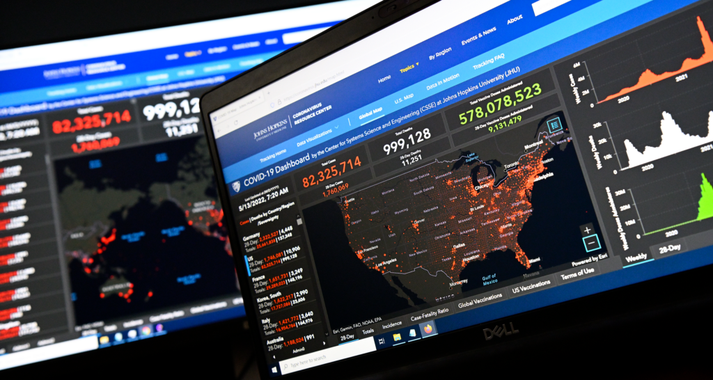 Two maps - one of the world, one of the U.S. - are shown on adjacent computer screens, with red dots tracking the spread of COVID-19 cases