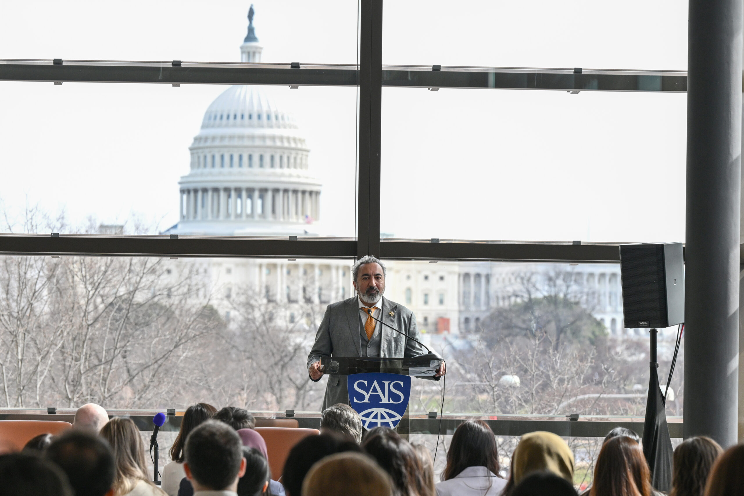 U.S. Rep. Ami Bera speaks to a crowd from a podium bearing a blue SAIS logo; the U.S. Capitol dome is visible through the large glass wall behind him