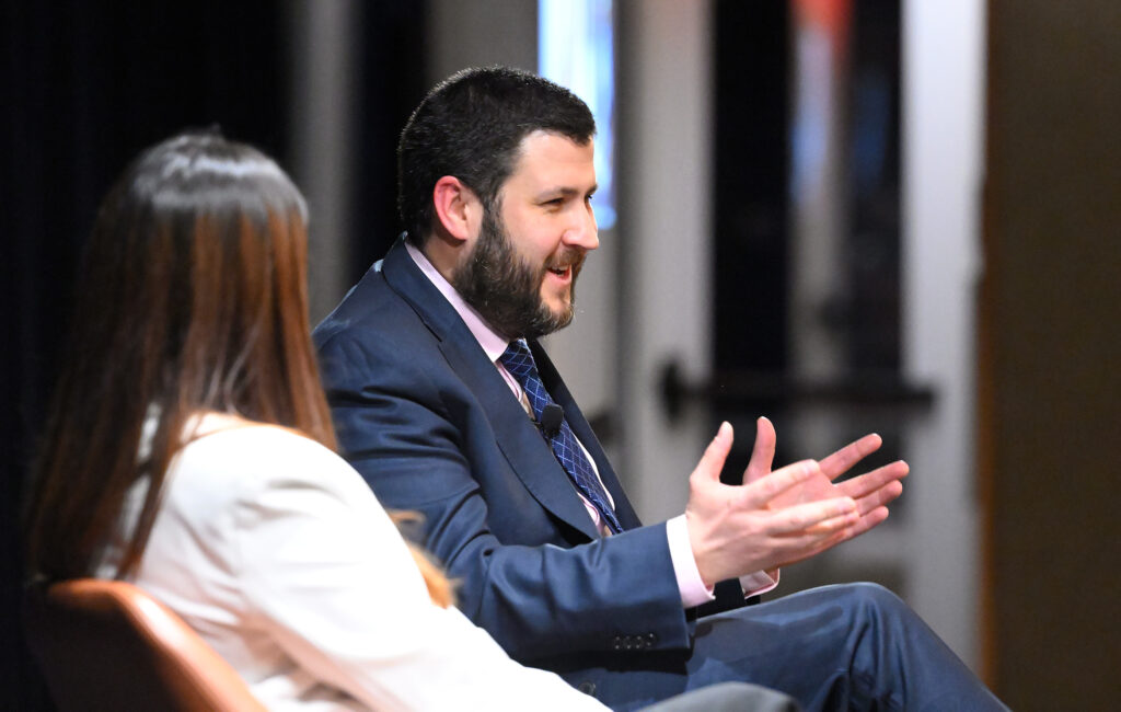 A bearded man in a gray suit speaks while seated next to a woman in a cream-coloreds top